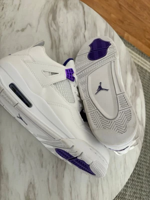 Vita sneakers med lila detaljer - Snygga vita sneakers från Jordan med lila detaljer på snörningen och sulan. Skorna har en klassisk siluett med meshpaneler och synlig Air-enhet i sulan. Perfekta för dig som vill sticka ut med stilrena och sportiga skor.