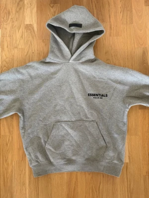Grå Essentials hoodie med tryck - Snygg grå hoodie från Essentials med svart tryck på bröstet. Klassisk känguruficka framtill och stor huva. Mjuk insida för extra komfort, perfekt för en chill och avslappnad stil. Size XS men sitter precis som en S eftersom essentials kläder är oversized. Den är använd en gång men har kvar tagsen hemma. Plagget är 100% äkta.