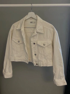 Vit cropped jeansjacka - Snygg vit jeansjacka i cropped modell med klassisk krage och knappstängning framtill. Två bröstfickor med lock och silverfärgade knappar ger en trendig look. Perfekt att slänga över en t-shirt för en cool och avslappnad stil.