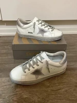 Golden Goose skor - Snygga vita sneakers med gråa skosnören och en silverfärgad stjärna på sidan. Skorna har silvriga detaljer vid häl och plös samt en mönstrad sula. Tillverkade i läder med en klassisk låg profil och trendig design.