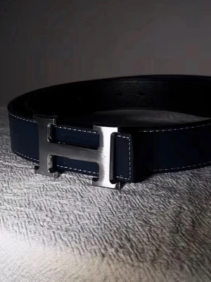 H bälte - Snyggt svart skärp med ett stort, silverfärgat H-format spänne. Skärpet har vita kontrastsömmar längs kanterna och är gjort i slätt skinn. Perfekt accessoar för att lyfta din outfit.