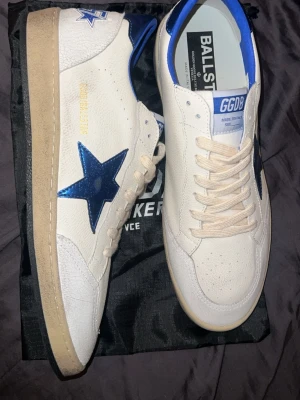 Vita sneakers med blå stjärna - Snygga vita sneakers från Golden Goose med blå metallicstjärna på sidan och blå detaljer vid hälen. Skorna har beige sula, vita skosnören och GGDB-logga på plösen. Ovandelen är i läder med perforerade detaljer framtill för extra ventilation.