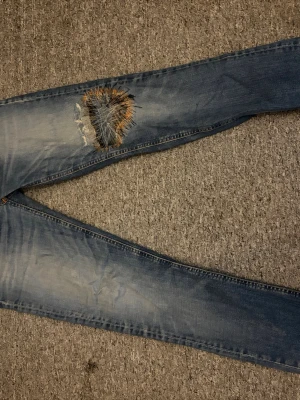 Ljusblå jeans med broderad lagning - Säljer ett par ljusblå jeans med unik broderad lagning på ena benet. Jeansen har en smal passform och klassisk femficksdesign. Materialet är bomull och de har slitningar för en avslappnad look. W 31 L 32