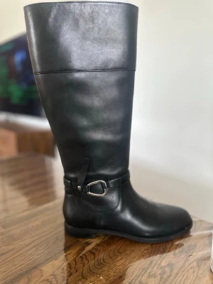 Svarta knähöga läderstövlar - Stilrena svarta knähöga stövlar i slätt läder med dekorativt spänne vid ankeln. Klassisk rund tå och låg klack gör dem perfekta till många outfits. Dragkedja på insidan för enkel på- och avtagning. Snygg och tidlös modell för dig som gillar boots med attityd. Orginalpriset ligger på 3000