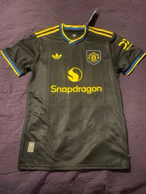 Svart Manchester United matchtröja - Snygg svart Manchester United matchtröja från Adidas med gula och blå detaljer. Tröjan har klubbmärke och sponsorlogga på bröstet samt gula ränder på axlarna. Tillverkad i lätt och ventilerande material, perfekt för fotbollsträning eller match. Inge namn. Pris kan sänkas vid snabb affär