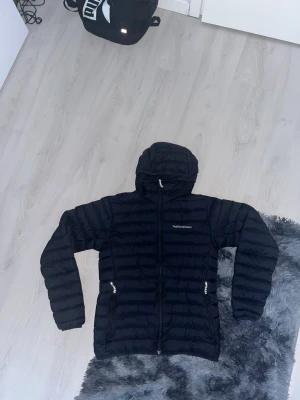 Svart pufferjacka med huva - Snygg svart pufferjacka från peak performance med huva och dragkedja framtill. Jackan har två dragkedjeförsedda fickor på sidorna och quiltad design som ger en sportig look. Perfekt för kalla dagar och enkel att matcha med olika outfits.