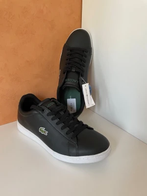 Svarta Lacoste sneakers i läder, stl 41, nya oanvända  - Snygga svarta sneakers från Lacoste i slätt läder med klassisk krokodillogo på sidan. Skorna har svarta skosnören, vit sula och diskret Lacoste-märke på plösen. Perfekta för en clean och stilren look. stl 41, nya oanvända 