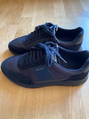 Svarta sneakers från Axel Arigato - Snygga svarta sneakers från Axel Arigato med klassisk siluett. Skorna har ovandel i textil och mocka, svarta skosnören och diskret logga på sidan. Perfekta för en clean och stilren look.