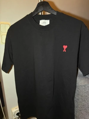 Ami Paris t shirt M - Svart t-shirt från Ami med ett litet rött hjärta broderat på bröstet. Klassisk rund hals och korta ärmar. Tillverkad i mjuk bomull som känns skön mot huden. Perfekt för en clean och stilren look. M form och sitter löst och bekvämt, helt ny utan prislapp