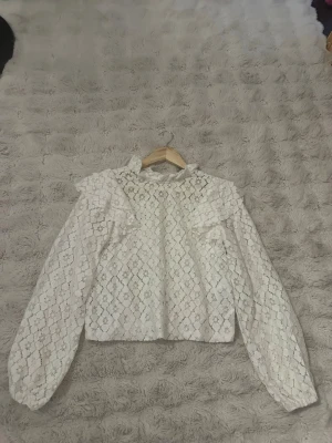 Vit spetsblus med volangdetaljer - Supersöt vit blus i spets med volangdetaljer vid axlarna och långärmad design. Blusen har ett romantiskt mönster och är lätt transparent tack vare det tunna spetsmaterialet. Perfekt för dig som gillar en feminin och trendig stil. Lösa trådar förekommer 