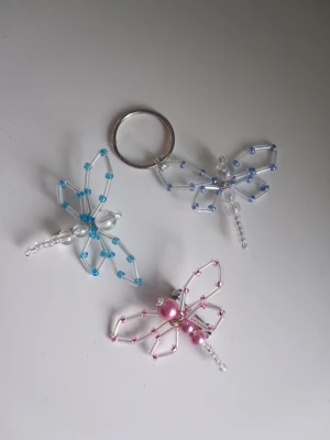 Nyckelring /brosch/pins trollsländor av pärlor 3st - Tre handgjorda trollsländor av pärlor i blått, rosa och skimmerfärgade pärlor. En av trollsländorna är fäst på en nyckelring. Och 2st är broscher/pins. Blir en söt liten accessoar till tex väskan. Mått ca 4x5cm