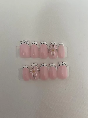 Press-on naglar rosa med prickig tipp - Säljer ett set press-on naglar i ljusrosa med vita tippar dekorerade med svarta prickar. Några naglar har även 3D-blommor med gulddetaljer för extra wow-effekt. Naglarna har en oval form och är gjorda i plast.