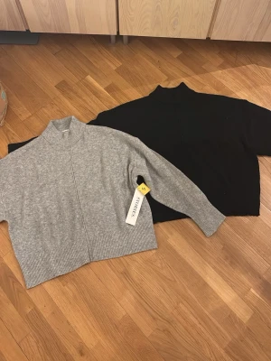 Grå och svart stickad tröja - Två stickade tröjor i croppad modell, en i ljusgrått och en i svart. Båda har hög krage och ribbstickade muddar vid ärmslut och nederkant. Perfekta att matcha med jeans eller kjol för en clean look.