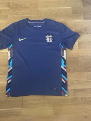 Blå England Nike fotbollströja - Snygg blå fotbollströja från Nike med Englands landslagsmärke på bröstet och gul Nike-logga. Tröjan har korta ärmar och färgglada detaljer i sidorna med inslag av vitt, blått och vinrött. Perfekt för match eller träning.