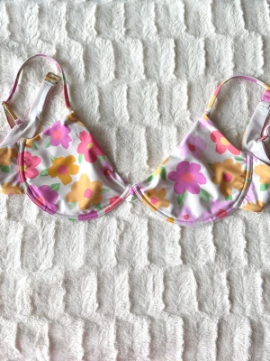 Blommig bikiniöverdel i pastellfärger - Söt bikiniöverdel med stora blommor i rosa, lila och gul på vit botten. Justerbara axelband och bygel för extra stöd. Perfekt för sommarens strandhäng eller poolparty.