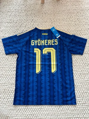 Blå Sverige tröja Adidas nr 17 gyökeres - Säljer en blå matchtröja från Adidas med Sveriges landslagsemblem och nummer 17 i gult. Tröjan har retroinspirerat mönster, vita och gula detaljer samt klassiska Adidas-ränder på axlarna. Perfekt för dig som älskar fotboll och vill sticka ut på läktaren.