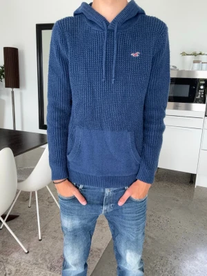 Blå stickad hoodie från hollister - Snygg blå stickad hoodie med luva och magficka. Tröjan har ett diskret märke broderat på bröstet och ribbade muddar vid ärmslut och nederkant. Perfekt för dig som gillar en avslappnad och stilren look. Tröjan är S och passar bäst runt 178-188
