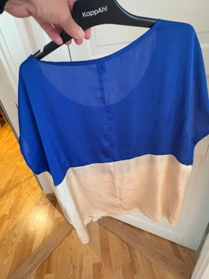 Blå och beige blus från KappAhl - Snygg tvåfärgad blus från KappAhl med blå överdel och beige nedtill. Blusen har lös passform, rund halsringning och korta ärmar. Materialet känns lätt och luftigt, perfekt för varmare dagar.