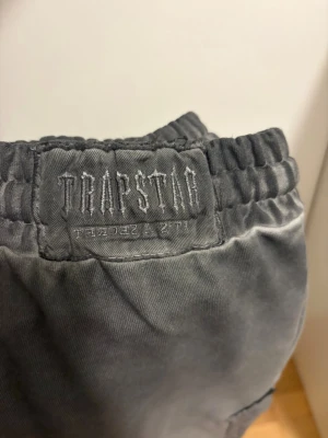 Grå mjukisbyxor från Trapstar - Säljer ett par grå mjukisbyxor från Trapstar med broderade detaljer och patchar på benen. Byxorna har resår i midjan med dragsko och är tillverkade i mjukt material för maximal komfort. Perfekta för en avslappnad och trendig look.
