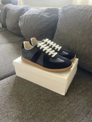 Maison Margiela Gats - Stilrena svarta sneakers med vita skosnören och beige innersula. Ovandelen är i en mix av mocka och slätt läder, och sulan är i klassisk gummi. Perfekta för en clean och tidlös look.