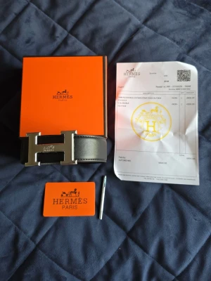 Herm3s svart skärp med H-spänne - Säljer ett svart skärp från Herm3s med det ikoniska H-spännet i silverfärgad metall. Skärpet är tillverkat i slätt skinn och levereras med originalbox och certifikat. Perfekt accessoar för att lyfta vilken outfit som helst.
