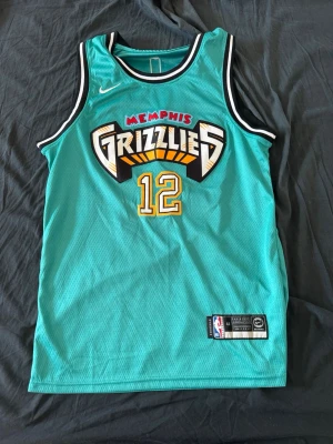 Memphis Grizzlies basketlinne #12 - Turkost basketlinne med Memphis Grizzlies-logga och nummer 12 i gult och vitt. På ryggen står det 'Morant' och siffran 12. Linnet har svarta och vita detaljer vid ärmhål och hals. Tillverkat i lätt och ventilerande meshmaterial.