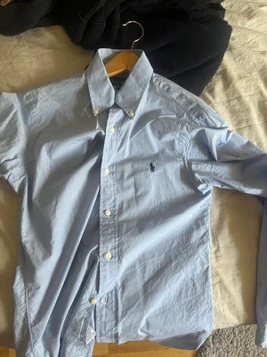 Ljusblå skjorta från Ralph Lauren - Klassisk ljusblå skjorta från Ralph Lauren i slim fit-modell. Skjortan har knappar framtill, button down-krage och den ikoniska broderade loggan på bröstet. Tillverkad i mjuk bomull och passar perfekt till både jeans och chinos.