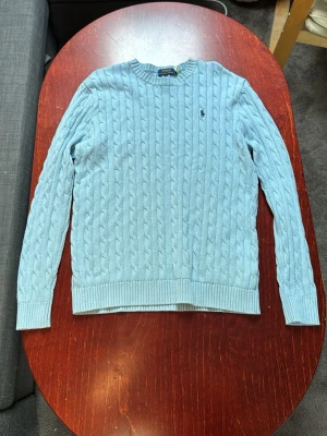 Ljusblå kabelstickad tröja Polo Ralph Lauren - Kabelstickad ljusblå tröja från Polo Ralph Lauren med rund halsringning och ribbade muddar. Tröjan har lång ärm och den klassiska broderade loggan på bröstet. Perfekt för en chill och stilren look. Storlek M