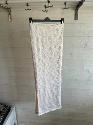 Vit långkjol med spets och blommönster - Säljer en elegant vit långkjol med blommigt spetsmönster. Kjolen har rak passform och är helfodrad, vilket ger en snygg och stilren look. Perfekt för dig som vill ha en romantisk och trendig kjol till sommaren eller speciella tillfällen.