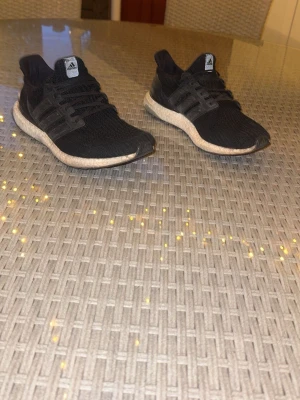 Svarta Adidas Ultraboost sneakers - Säljer ett par svarta Adidas Ultraboost sneakers med stickad ovandel och vita Boost-sulor. Skorna har klassiska tre ränder på sidan och svart yttersula med Continental-gummi för extra grepp. Perfekta för dig som gillar stilrena och bekväma sneakers.
