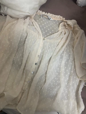 Beige transparent blus från Zara - Söt beige blus från Zara i transparent, prickigt tyg med volangkant vid halsen och knappar framtill. Blusen har korta ärmar och fina spetsdetaljer vid axlarna. Perfekt för en trendig och romantisk look.