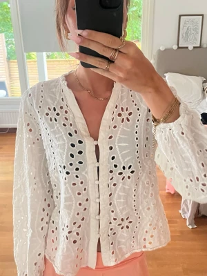 Vit broderad blus med knappar - Säljer en superfin vit blus med broderade hålmönster och klädda knappar framtill. Blusen har v-ringning och långa, luftiga ärmar. Materialet känns lätt och somrigt, perfekt till jeans eller kjol för en fräsch look.