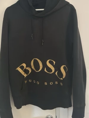 Svart hoodie från Hugo Boss - Säljer en svart hoodie från Hugo Boss med stor guldfärgad BOSS-logga och texten HUGO BOSS på magen. Hoodien har huva med dragsko och en klassisk känguruficka framtill. Perfekt för dig som gillar stilrena och exklusiva plagg. Den är lite solbränd men det är en unik design på hoddien som inte säljs längre.