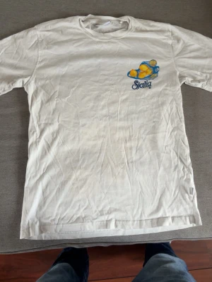 Vit Sicilia t-shirt med citrontryck - Säljer en vit t-shirt med färgglatt tryck av citroner i nät och texten 'Sicilia Italy' i blått och gult. T-shirten har rund hals och korta ärmar. Perfekt för dig som gillar unika motiv och vill sticka ut lite extra.