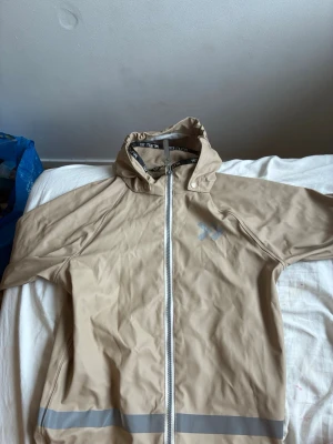 Beige regnjacka med huva och reflex - Snygg beige regnjacka med hel dragkedja framtill, huva och reflexdetaljer både fram och bak. Jackan har ett minimalistiskt tryck på bröstet och är tillverkad i vattenavvisande material. Perfekt för regniga dagar när du vill hålla dig torr och synlig.