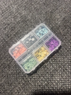 Nageldekorationer i mixade färger - Säljer ett set med små nageldekorationer i form av blommor i olika färger: rosa, blå, lila, gul och grön. Kommer i en praktisk plastlåda med sex fack. Perfekt för att skapa kreativa och färgglada nageldesigner.
