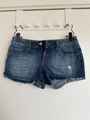 Blå jeansshorts från Esprit - Snygga blå jeansshorts från Esprit med råa kanter och slitningar för en avslappnad look. Klassisk femficksmodell med knapp och dragkedja framtill. Perfekta till sommaren och lätta att matcha med olika stilar.