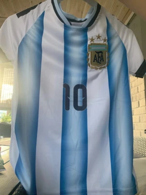 Argentina fotbollströja och shorts #10 - Säljer ett matchställ med Argentina fotbollströja och svarta shorts. Tröjan har blå och vita ränder, nummer 10 och AFA-emblem med tre stjärnor. Shortsen är svarta med nummer 10 och AFA-logga. Perfekt för dig som älskar fotboll och vill representera Argentina.