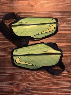 Nike benskydd Protegga Flex grön - Säljer ett par Nike Protegga Flex benskydd i ljusgrönt med svart kant och justerbara kardborreband. Skydden har en snygg design med Nike-logga och är tillverkade i plast och skum för bra komfort och skydd vid sportaktiviteter.