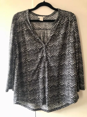 H&M Mönstrad Blus 3/4-ärm V-ringad – Stl S - Snygg och lättburen blus från H&M i svartvitt grafiskt mönster. Tillverkad i ett tunt, lätt genomskinligt tyg som faller fint. Modellen har 3/4-ärm, liten krage, V-ringning med tre knappar fram samt små slitsar i sidorna och är något längre baktill. Märke: H&M Storlek: S Mått: Axelbredd ca 38 cm, längd ca 65 cm Material: 100% Polyester Skick: Mycket gott skick