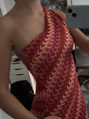 Mönstrad oneshoulder kortklänning - Säljer en kort, figurnära oneshoulder-klänning med färgglatt mönster i rött, rosa och vitt. Klänningen har ett diagonalt mönster och är ärmlös, perfekt för dig som vill sticka ut. Materialet känns mjukt och stretchigt.
