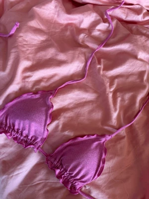 Rosa glittrig trekantsbikini - Supersöt rosa trekantsbikini med glittrigt tyg och vågiga kanter. Bikinin har knytband i nacken och ryggen, perfekt för soliga dagar på stranden eller poolen. Har köpt på mango teen i Spanien. Har även underdelen till som jag kan skicka bild på.