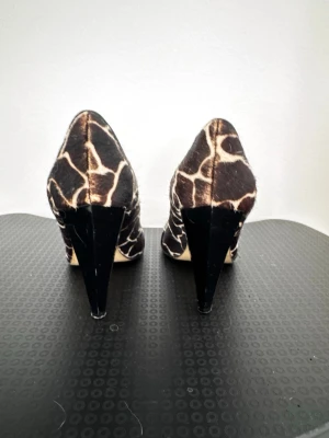 Bruna pumps med giraffmönster - Snygga pumps med giraffmönstrad ovandel i brunt och beige. Skorna har öppen tå och svarta lackdetaljer framtill. Klackarna är höga och smala, vilket ger en elegant siluett. Perfekta för dig som vill sticka ut med ett djurmönstrat statement.