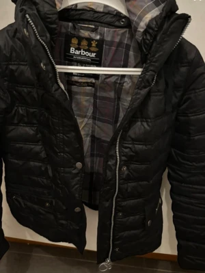 Svart pufferjacka från Barbour - Snygg svart pufferjacka från Barbour med quiltad design och dragkedja framtill. Jackan har tryckknappar, hög krage och ett klassiskt rutigt innerfoder. Perfekt för kalla vinterdagar och riktigt stilren look.