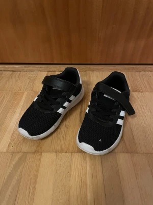 Svarta adidas sneakers  - Snygga svarta sneakers från adidas med vita ränder på sidorna och vit sula. Ovandelen är i meshmaterial för bra andningsförmåga och skorna har både kardborreband och svarta skosnören. Perfekta för en sportig och avslappnad stil.