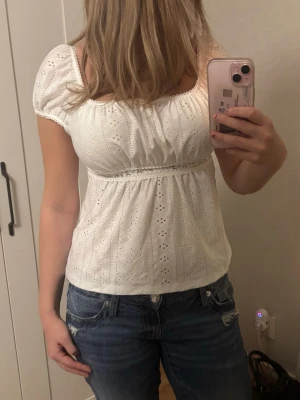 Vit broderad blus med puffärm - Söt vit blus med broderade detaljer och puffärmar. Toppen har en smickrande skärning under bysten och är tillverkad i bomull. Perfekt för dig som gillar romantisk och feminin stil.
