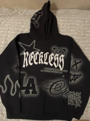 Svart Reckles hoodie med graffiti-tryck - Säljer en svart hoodie med streetwear-vibe och stora graffiti-inspirerade tryck i vitt och grått. Framsidan har texten 'RECKLESS', LA-logga, björnmotiv och flera coola detaljer som stjärnor och eldflammor. Oversized passform och mjukt material.
