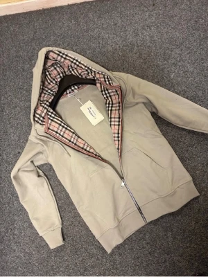 Beige hoodie med dragkedja från Burberry - Snygg beige hoodie från Burberry med dragkedja och klassiskt rutigt foder i huvan. Broderad Burberry-logga på bröstet och två stora fickor framtill. Tillverkad i mjukt bomullsmaterial och har ribbade muddar vid ärmslut och nederkant.