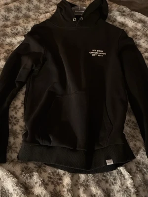 Svart hoodie Les Deux Copenhagen - Svart hoodie från Les Deux Copenhagen med vit text på bröstet och klassisk känguruficka. Huva med dragsko och ribbade muddar vid ärmslut och nederkant. Perfekt för en avslappnad och stilren look.