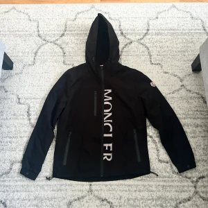 Svart vindjacka från Moncler med huva - Svart vindjacka från Moncler med stor vit logga framtill, hel dragkedja och huva. Jackan har flera praktiska fickor med dragkedja och justerbar nederkant. Perfekt för dig som vill ha en clean och sportig look med premiumkänsla.